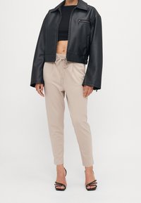 Czarna kurtka bomber z sztucznej skóry z kieszonią na zamek, zestawiona z beżowymi spodniami typu tapered i czarnymi sandałami na paski.
