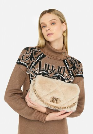 Bolso de piel beige con diseño acolchado, cadena dorada como correa y cierre con logo, sostenido por una modelo con suéter estampado.