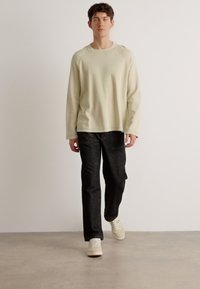 Light beige long-sleeve shirt with a textured fabric, paired with dark gray pants and cream sneakers, standing on a neutral background.