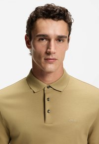 Beżowa koszulka polo z krótkim rękawem, wykonana z gładkiej tkaniny, z klasycznym kołnierzykiem, trzema guzikami oraz haftowanym logo "BOSS" na piersi.