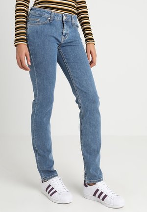 Personne portant un jean bleu droit, des baskets blanches avec des rayures violettes et un haut à manches longues rayé noir, jaune et blanc.