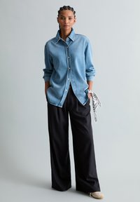 Frau steht mit Händen in den Taschen, trägt hellblaues Jeanshemd, schwarze weite Hosen, beigefarbene Schuhe und ein gemustertes Tuch, das am Handgelenk gebunden ist.