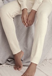 Crèmekleurige, zachte gebreide loungewear met geribbelde manchetten, gecombineerd met blote voeten die op een grijze visgraatpatroondeken rusten.