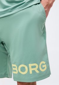 Ανοιχτό πράσινο σορτς με πλαϊνή τσέπη και μεγάλα διάτρητα κίτρινα γράμματα "BORG" κοντά στο τελείωμα, φορεμένο με αντίστοιχο πουκάμισο.