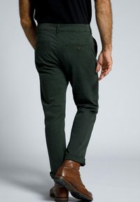 Pantalones de algodón verde con corte ajustado, dos bolsillos traseros y detalles de botones marrones. Llevados con botas de cuero marrones.