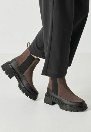 Persona che indossa stivaletti Chelsea marroni in camoscio e neri robusti con lati elastici neri e pantaloni neri su uno sfondo chiaro uniforme.