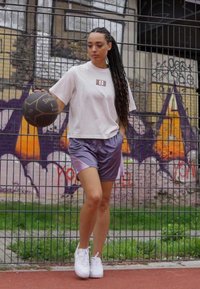 Witte t-shirt met korte mouwen en een klein logo, gecombineerd met paarse sportshorts. Witte sneakers en houdt een zwarte basketbal vast. Achtergrond van een basketbalveld.