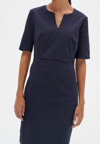 Mörkblå klänning med en figursydd silhuett, korta ärmar och v-ringning. Tillverkad av ett slätt tyg med subtila sömnadsdetaljer.