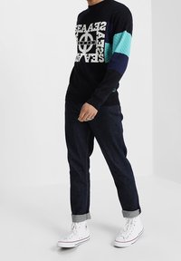 Schwarzer Strickpullover mit geometrischen blauen und türkisfarbenen Ärmeln, versehen mit fettem weißen Text. Kombiniert mit dunklen Jeans und weißen High-Top-Sneakern.