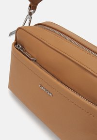 Sac zippé en simili cuir marron avec quincaillerie argentée, doté d'une poche avant et d'un logo en relief pour l'identification de la marque. Design compact.