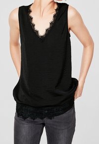 Černý top bez rukávů s véčkovým výstřihem a krajkovým lemem. Obsahuje strukturovanou tkaninu a vrstvený vzhled s krajkou na spodní části. Nosič má na sobě šedé kalhoty.
