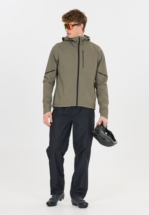 Mand iført olivengrøn jakke, sorte bukser, cykelsko og orangefarvede solbriller, holder en sort cykelhjelm i højre hånd.