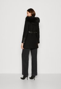 Cappotto nero con cappuccio in pelliccia sintetica, dettagli in pelle sulle maniche e vita cintrata. La modella indossa jeans scuri a gamba larga e décolleté neri.