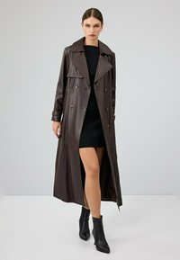 Bruine leren trenchcoat met een dubbelzijdig ontwerp, grote kraag en een zijsplit, gecombineerd met een aansluitende zwarte jurk en enkellaarzen.