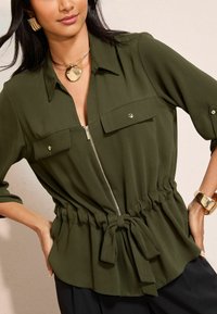 Sieviete, tērpusies olīvzaļā ar rāvējslēdzēju, ar jostu viduklī, zelta izteiksmīgām kaklarotām, masīvām zelta auskariem un atbilstošu aproci, rokas uz gurniem.