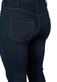 Jeans de mezclilla azul oscuro con un diseño ajustado, que presentan costuras naranjas y dos bolsillos traseros. Textura suave con detalles mínimos.