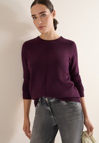 Fioletowy sweter dzianinowy z okrągłym dekoltem i ściągaczami na rękawach, w połączeniu z szarymi dżinsami. Gładka faktura; minimalistyczny design.