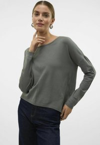 Pilkai megztinis su apvalia kaklo iškirpte ir nuleistomis pečių siūlėmis, turintis glotnią tekstūrą ir laisvą kirpimą, derinamas su tamsiai mėlynomis džinsomis.