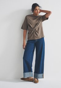 Brown knit top with a knotted front, paired with wide-leg blue denim jeans featuring a rolled cuff. Brown flats complete the look.