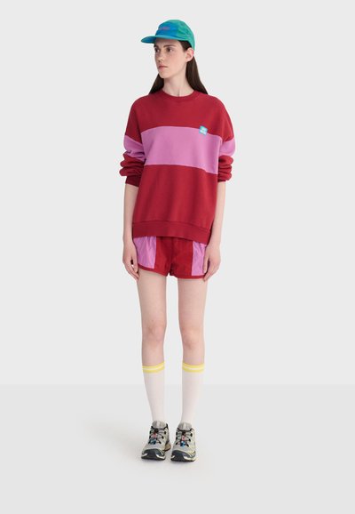 Jeune personne portant un sweat-shirt à blocs de couleurs rouge et rose ainsi que des shorts, des chaussettes blanches montantes avec des rayures jaunes, des baskets grises et une casquette vert-bleu.