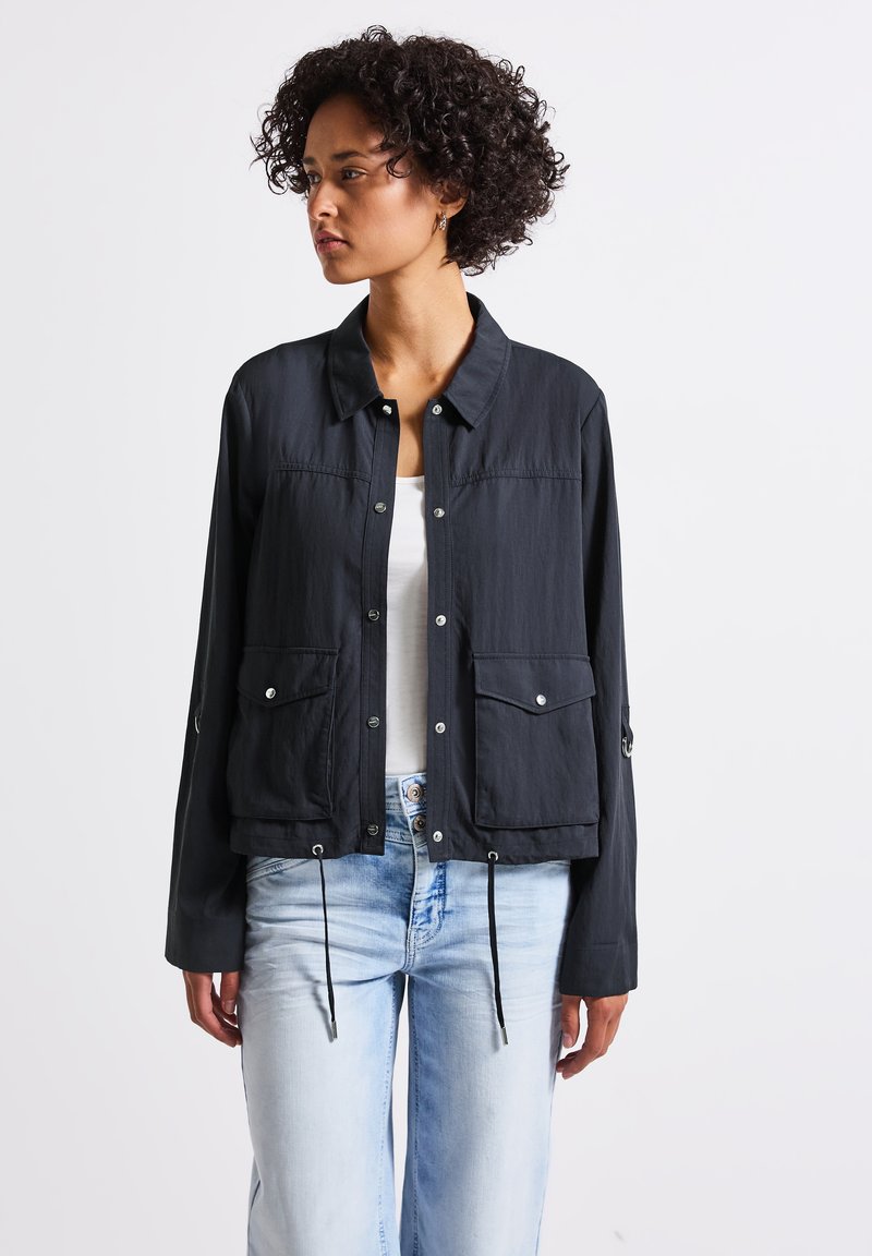 Modèle avec des cheveux bouclés portant une veste noire par-dessus un haut blanc, associée à un jean bleu clair. L'accent est mis sur sa tenue élégante.