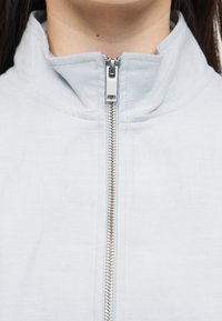 Gros plan sur le col d'une veste gris clair à fermeture éclair remontée jusqu'en haut, montrant la fermeture éclair en métal et le tissu texturé.