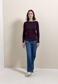 Długie rękawy, prążkowana bluzka w ciemnofioletowym kolorze, noszona z jasnoniebieskimi dżinsami w fasonie dzwonów i białymi sneakersami, stojąca na neutralnym tle.