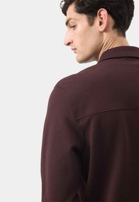 Mørk burgunder skjorte med struktureret stof, der har en krave og synlige syninger langs skuldrene og ryggen.