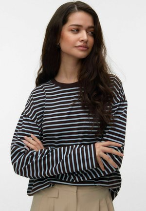 Młoda kobieta o długich ciemnych włosach, ubrana w brązowo-niebieską koszulę w paski z długim rękawem i beżowe spodnie, z założonymi rękami, patrzy na bok.