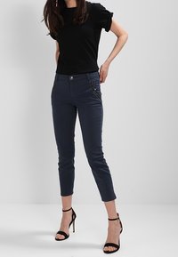 Černý top s krátkým rukávem, námořnické slim-fit kalhoty s kapsami na zip a černé sandály na vysokém podpatku. Hladká tkanina s neformálním, moderním designem.
