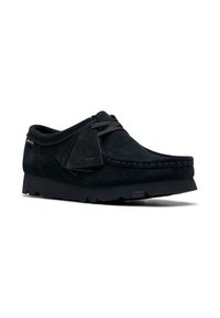 Sort suede mokasino sko med snøre og kvastdetalje, tyk sål og syninger langs tåen, vinklet for at vise ydersiden og forsiden.