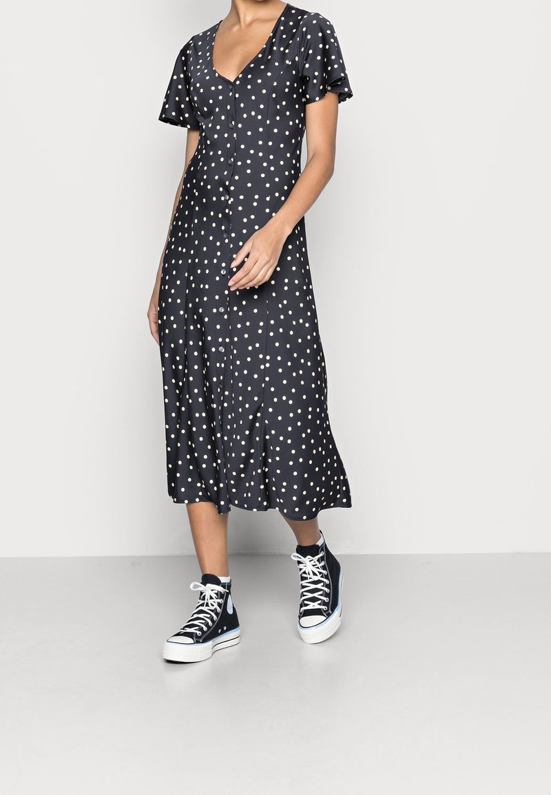 Femme portant une robe midi noire à pois blancs avec des boutons sur le devant, associée à des baskets montantes noires avec des lacets blancs.