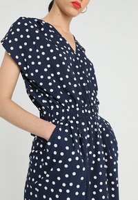 Marineblauer Pünktchen-Jumpsuit mit kurzen Ärmeln, V-Ausschnitt, elastischem Taillenbund und Seitentaschen, verziert mit großen weißen kreisförmigen Mustern.