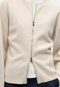 Cardigan beige en tissu doux, avec une fermeture éclair à l'avant, un col rond et des manches longues. Texture lisse avec une coupe décontractée.