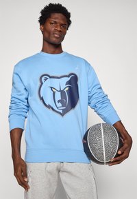 Világoskék pulóver, elöl szürke és sötétkék medve grafikával, bordázott mandzsettákkal és aljjal, szürke melegítőnadrággal és egy kosárlabdával párosítva.