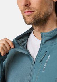 Vaaleansininen vetoketjullinen takki, jossa on teksturoitu sisävuori. Ominaisuuksiin kuuluu korkea kaulus, vetoketjukiinnitys ja "SWEDEMOUNT" -logo rinnassa.