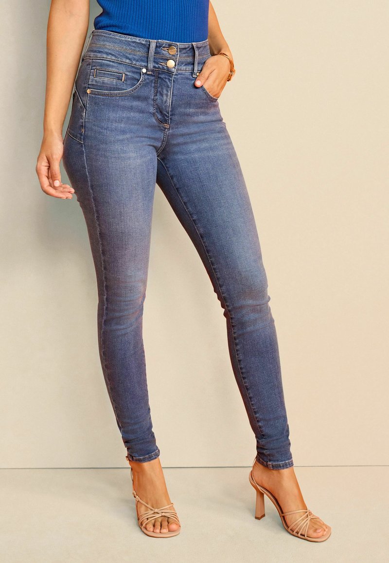 Jeans ajustados en denim azul oscuro con un acabado ligeramente desgastado. Presentan una cintura alta, dos bolsillos frontales y detalles en oro.