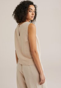 Haut sans manches, couleur beige clair, avec un col rond, un détail de dos en forme de serrure, et une texture lisse. Associé à un pantalon léger assorti.