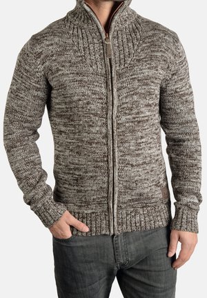 Man draagt een bruine en grijs gemêleerde gebreide trui met rits, met de handen gedeeltelijk in de zakken, gecombineerd met donkergrijze spijkerbroek.