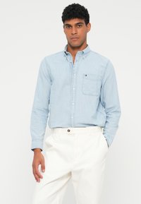 Camisa azul claro de botão, em algodão, com um bolso no peito, combinada com calças brancas. Apresenta um pequeno logotipo no bolso.