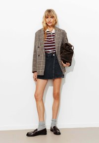 Blazer croisé en tartan, chemise rayée bordeaux et blanche, jupe en denim noire, chaussettes grises et mocassins marron, tenant un sac marron foncé.