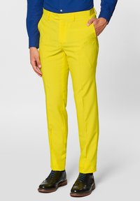 Pantaloni gialli su misura in un tessuto liscio, caratterizzati da una vestibilità slim e tasche laterali, abbinati a scarpe nere e a una camicia blu con bottoni.