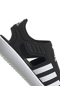 Svart atletisk sandaler med åpen side og hvit tre-stripe design, laget av mykt materiale, med en teksturert hvit såle.