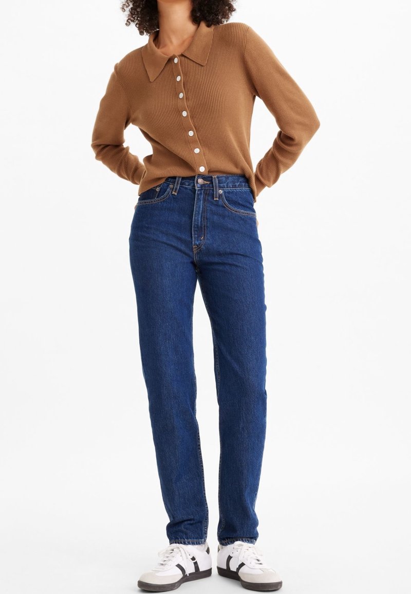 Kvinna i en brun knappad långärmad tröja, mörkblå högmidjade jeans och vita sneakers med svarta ränder.