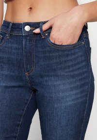 Primer plano de una persona que lleva pantalones vaqueros de mezclilla azul oscuro con costuras naranjas, con la mano metida en el bolsillo delantero.