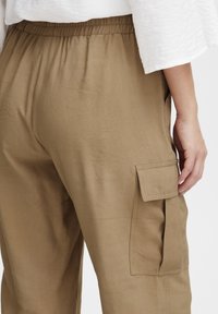 Bruna cargo byxor med elastiskt midjeband, sidofickor och mjuk textur, med en synlig cargoficka på vänster sida.