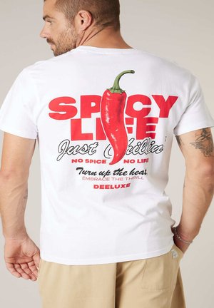 Homme portant un t-shirt blanc avec un grand motif de piment rouge et le texte « Vie épicée, juste chill, pas d'épices pas de vie ! » au dos.