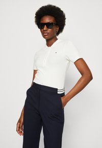 Polo bianco a maniche corte con colletto e un piccolo logo, abbinato a pantaloni navy con un accento a righe sulla vita. Occhiali da sole neri.