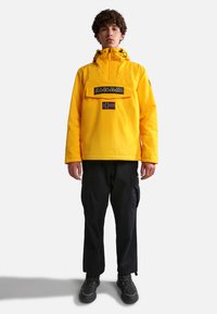 Yellow waterproof jacket with a front pocket, logo patch, and Norwegian flag. Worn over black cargo pants and black footwear.