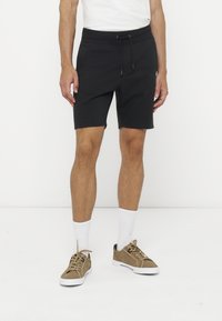 Mann trägt schwarze Shorts mit Kordelzug, weiße Sneakersocken und braune Turnschuhe und steht vor einem weißen Hintergrund.